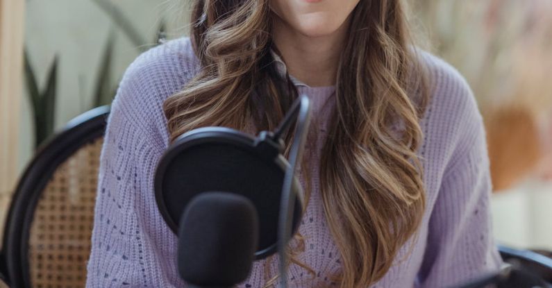 Hosting - Cheerful female radio host looking at camera while sitting in chair with opened notebook near professional microphone in light room